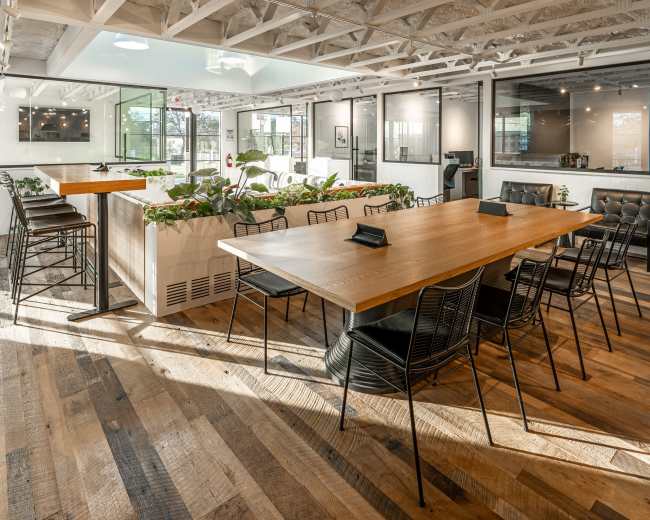 The image shows a spacious office interior featuring a large wooden table surrounded by black chairs, plants, and exposed ceiling beams.