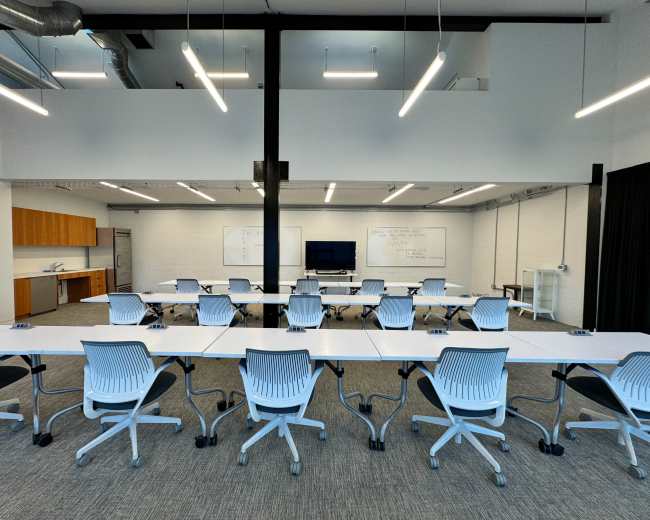 A modern classroom features rows of white tables with blue chairs facing a screen and whiteboards in a well-lit space.