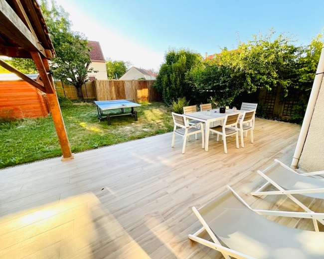 A patio with a dining table and chairs overlooks a grassy yard and a ping pong table surrounded by wooden fences and trees.