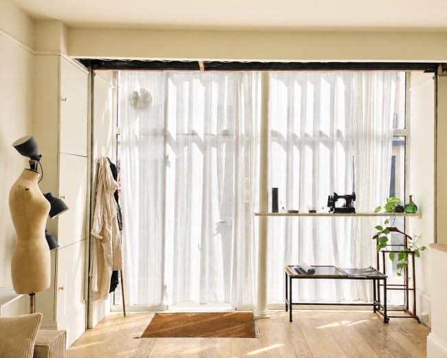 The image shows a well-lit interior space featuring sheer curtains over a window, a mannequin, a small table with a sewing machine, and a potted plant.