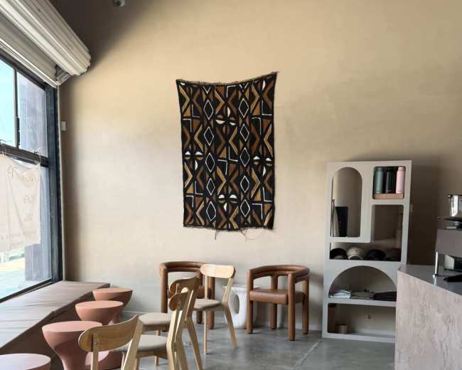 The image shows a modern café interior featuring a row of wooden chairs, small circular tables, a patterned wall hanging, and a minimalist shelving unit.