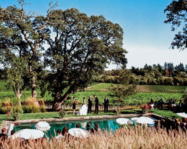 A wedding ceremony is taking place by a poolside in a vineyard, surrounded by large trees and guests seated under umbrellas.