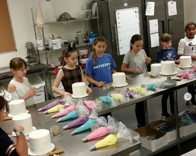 A group of children is decorating white cakes with colorful icing in a kitchen setting.