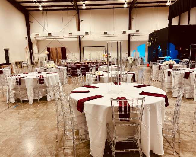 The image shows an indoor event space arranged with multiple tables covered in white tablecloths and set with clear chairs, featuring centerpieces and napkins in a deep red color.