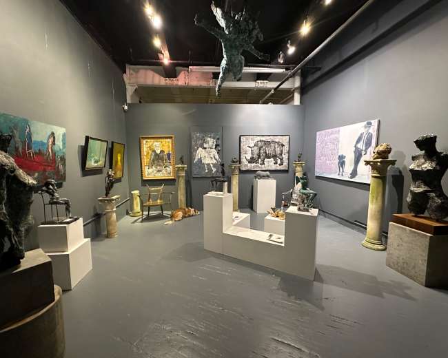 The image shows an art gallery featuring various sculptures and paintings displayed on gray walls and a gray floor, with a dog lying on the floor.