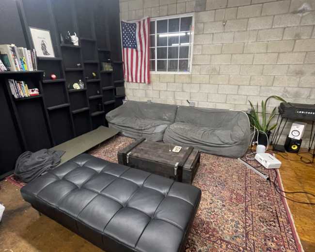 A minimalist living area features a gray sofa and a black ottoman on a patterned rug, with a shelf of books and an American flag on the wall.