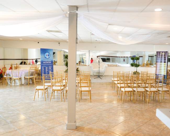 A spacious event venue is set up with rows of gold chiavari chairs and decorative drapery, ready for a gathering.