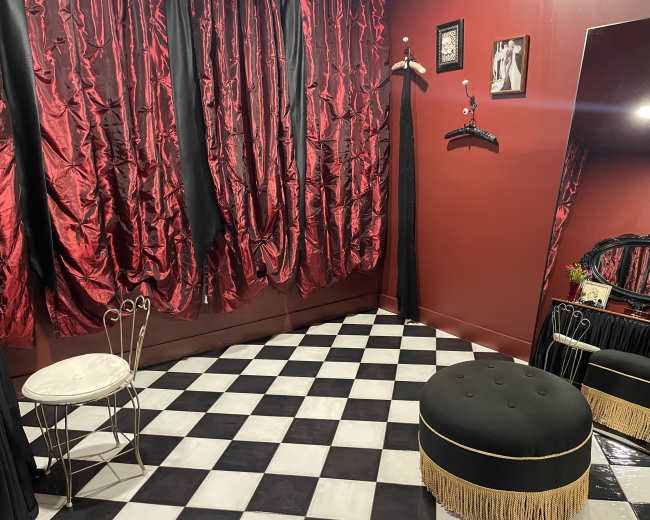 The image shows a room with red curtains, a black and white checkered floor, a round upholstered stool, a small chair, and a large decorative mirror.
