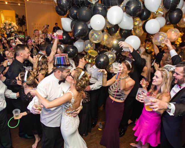 A large crowd celebrates at a festive event with balloons overhead and guests holding drinks while a couple dances in the center.