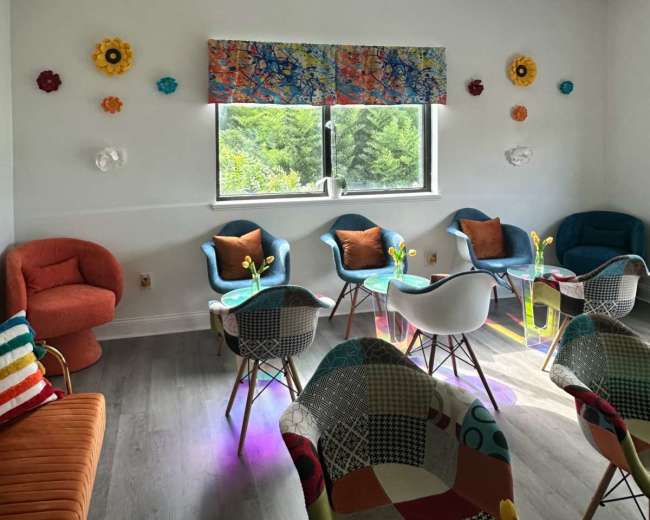 The image shows a brightly colored lounge area with various chairs, tables, and decorative flowers on the walls near two large windows.