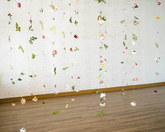 A collection of suspended flowers and leaves hangs from the ceiling in a bright, empty room with a white brick wall and wooden floor.