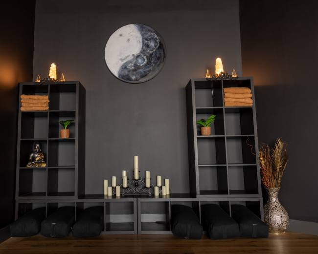 A serene room arranged with two dark shelving units, neatly stacked towels, a Buddha statue, candles, and plants, beneath a circular yin and yang wall art.