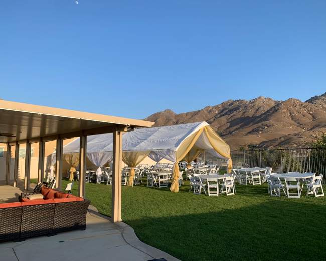 A tented outdoor event space with white tables and chairs is set up on green grass beside a building, with mountains in the background under a clear blue sky.