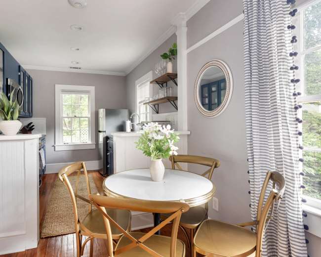 The image shows a modern kitchen with blue cabinetry, a circular white dining table surrounded by gold chairs, and large windows allowing natural light to enter.