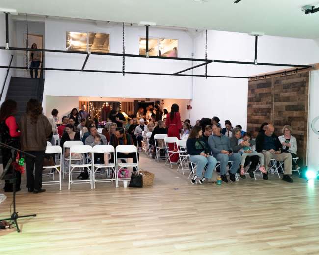 A large indoor space is filled with rows of white chairs, where a diverse audience gathers for an event, with a Christmas tree visible in the foreground.