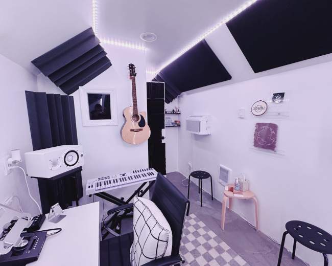 A small music studio with a guitar hanging on the wall, a keyboard on a desk, and acoustic panels on the ceiling.