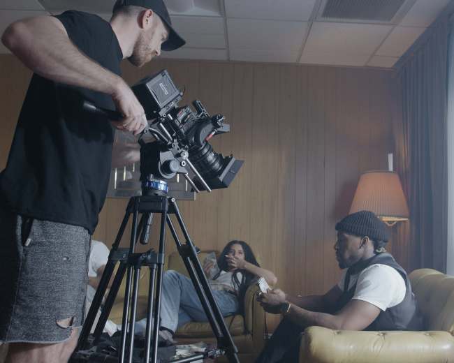 A person operates a camera on a tripod while another individual sits on a couch, looking at their phone, in a room with yellow lighting.
