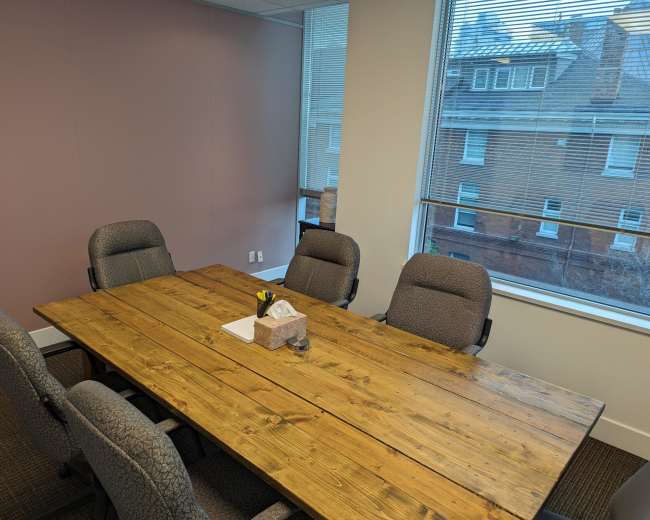 A rectangular wooden table is set with a notepad and a small decorative item in a conference room with gray chairs and large windows.