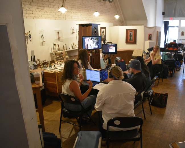 A group of people is working in a studio, seated around tables with computers and equipment, while others are positioned in the background among various artworks and supplies.