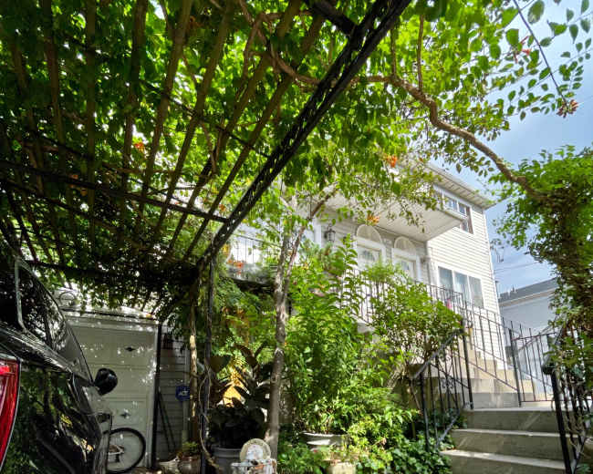 A car is parked under a trellis covered with vines, leading to a yard with steps and a well-maintained garden beside a house.