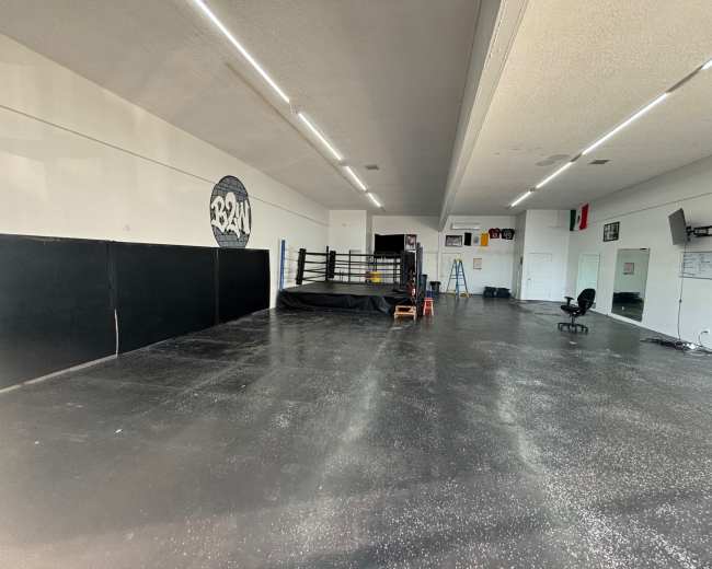 The image shows a spacious, empty boxing gym with a central ring, black padded walls, and minimal equipment scattered around.