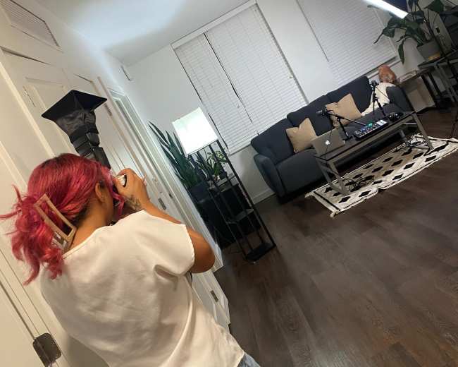 A person with pink hair takes a photo in a well-lit living room featuring a sofa, coffee table, and plants.