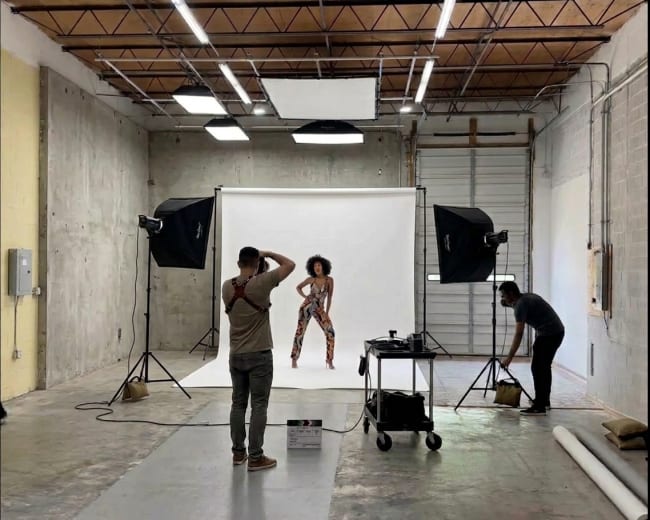 A model poses in front of a white backdrop while two photographers adjust equipment in a studio setting.