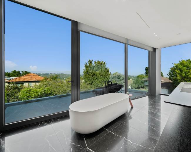 A modern bathroom features a freestanding tub and large windows that showcase a view of greenery and distant rooftops.