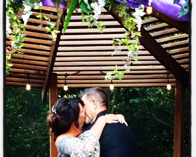 A couple kisses under a decorated gazebo with hanging flowers and string lights.