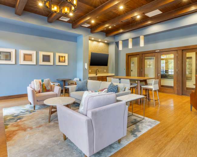The image shows a modern lobby area featuring comfortable seating, tables, and a wall-mounted television, set against a backdrop of blue and wooden tones.