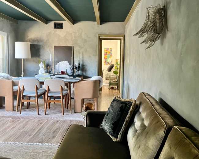 A cozy living room features a green ceiling, a beige sofa, a dining area with a round table, and decorative wall elements.