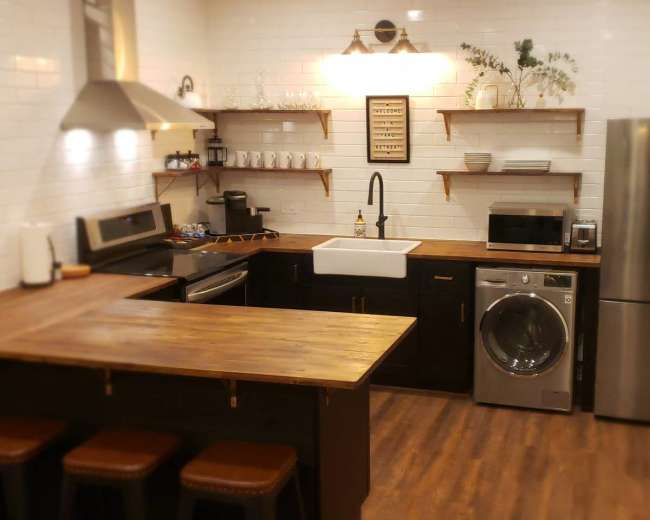 The image shows a modern kitchen with wooden countertops, black cabinets, stainless steel appliances, and open shelving.