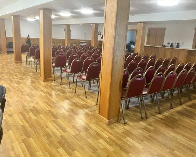 The image shows a spacious indoor hall with rows of red chairs set up neatly on a wooden floor, separated by wooden columns.