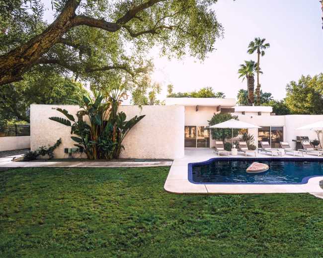 A modern house with large windows is situated by a swimming pool surrounded by lush greenery and palm trees.