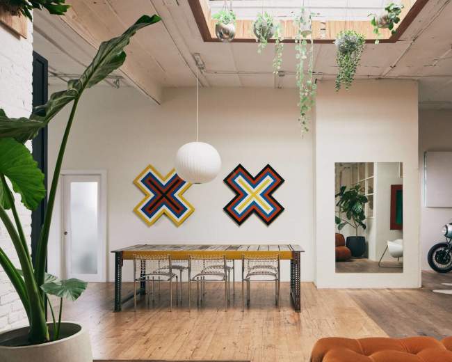 The image features a spacious room with a wooden dining table surrounded by chairs, two colorful geometric artworks on the walls, a large mirror, and a motorcycle beside a whiteboard.