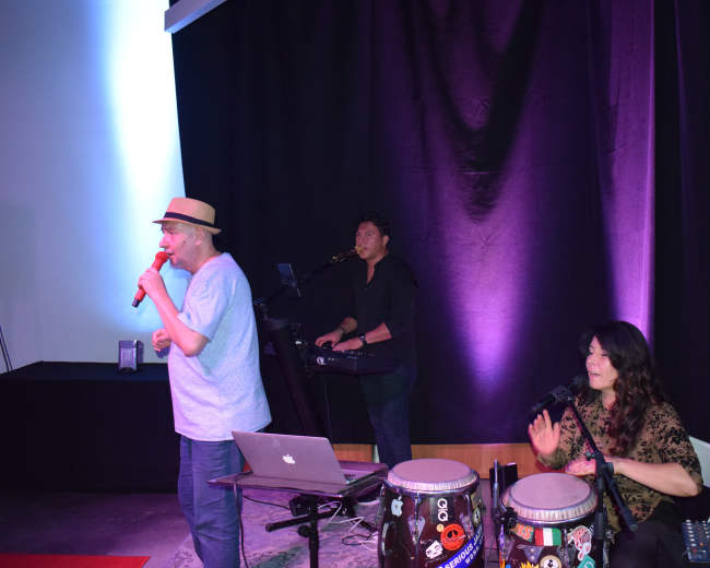 A man sings into a microphone while a woman plays conga drums and a man operates a keyboard behind them, set against a backdrop of purple lighting.