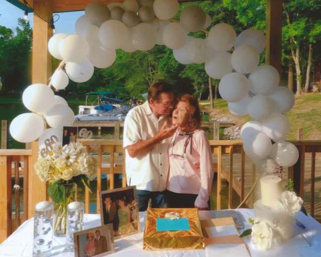 A couple celebrates a milestone anniversary under a balloon arch, surrounded by a decorated table with flowers, photos, and a cake by a lakeside.
