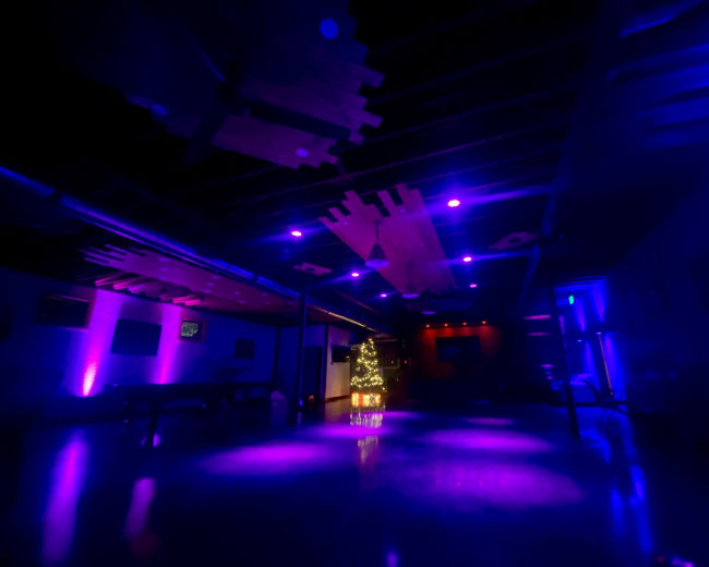 The image shows a dimly lit indoor space with purple lighting and a small, decorated Christmas tree in the corner.