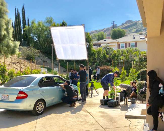 A film crew sets up equipment around a parked car in a garden area with hills in the background.