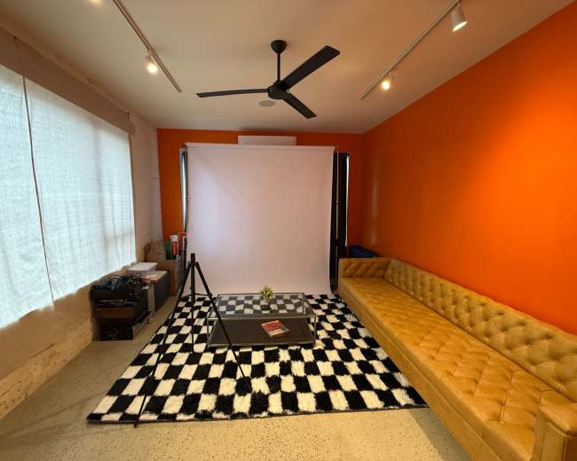 The image shows a modern interior space with an orange accent wall, a yellow tufted sofa, a black and white checkered rug, and a backdrop on a stand set up for photography.