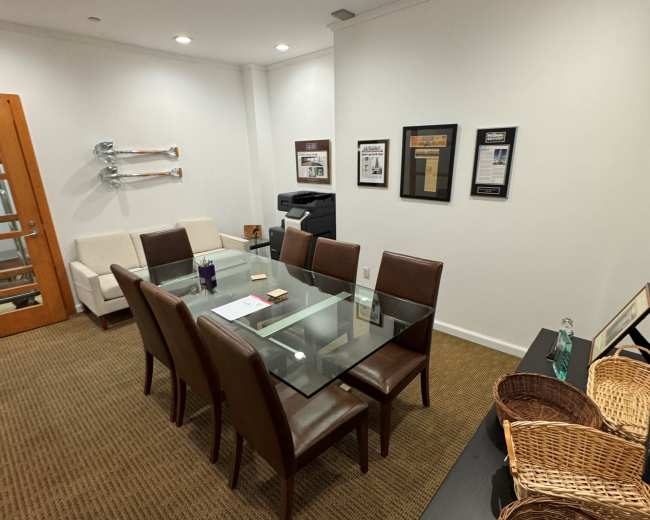 The image shows a conference room with a glass dining table surrounded by brown leather chairs, a sofa in the corner, and various framed documents on the walls.