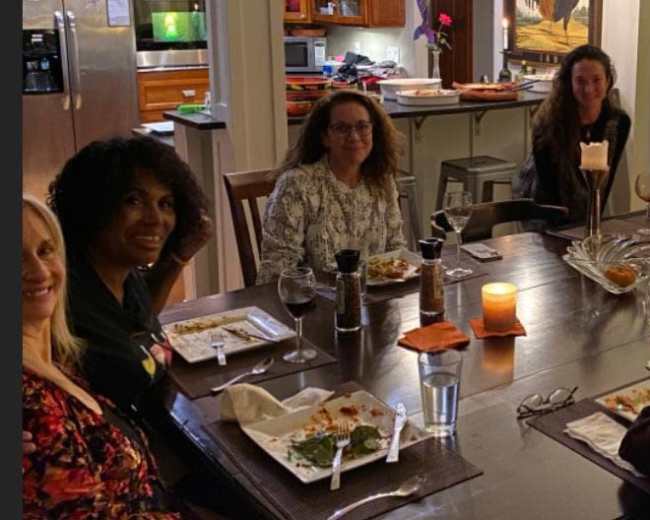 A group of eight women sit around a dining table with plates of food and drinks, surrounded by a warmly lit kitchen and living area.