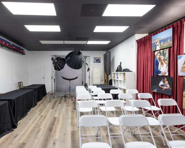 The image shows a spacious photography studio with two tables draped in black cloth on the left, a backdrop setup in the center, and rows of white chairs arranged in front of a red curtain showcasing framed photographs on the right.
