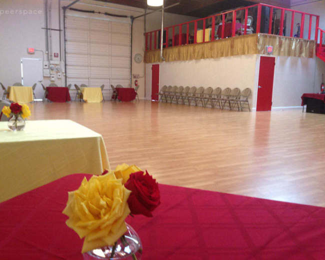 A spacious event hall features neatly arranged tables with yellow and red tablecloths and floral centerpieces, with a seating area along the wall.