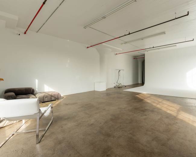 The image shows a spacious, empty studio with a white backdrop, concrete flooring, and minimal furniture including a couch and a stand.