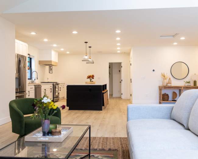 The image shows a modern living space featuring a light-colored sofa, a glass coffee table, and a view of a kitchen area with black cabinets and white countertops.