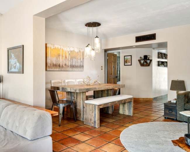 The image shows a bright, open living space featuring a dining area with a wooden table and benches, adjacent to a cozy seating area with a sofa.