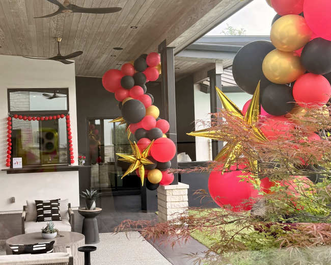 The image shows a covered outdoor space decorated with large clusters of balloons in red, black, and gold, along with star-shaped ornaments.