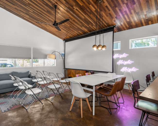 The image shows a modern meeting space with a mix of seating arrangements, including folding chairs and a long table, under a wooden ceiling.