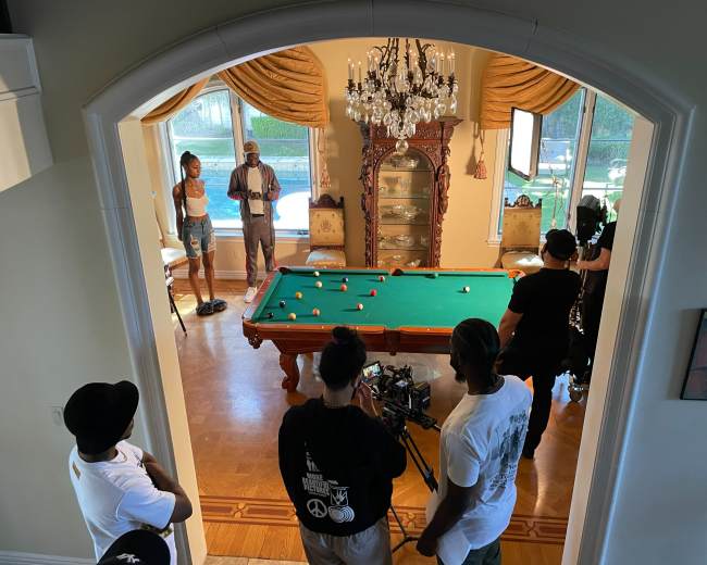 A film crew is shooting a scene involving a pool table in a well-lit room with large windows and elegant furniture.
